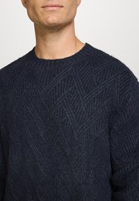 Marineblauw gebreid sweater met een diamanten textuurpatroon, ronde halslijn en zacht, dik materiaal. Close-up van de bovenkant van het lichaam.