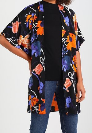 Nainen, jolla on musta, avoimella edestä oleva kimono, jossa oranssin, violetin ja pinkin kukkakuvio, mustan topin ja sinisten farkkujen päällä.