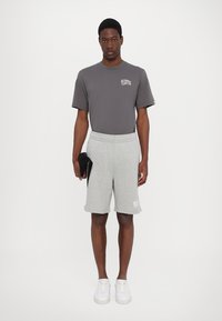 Grå t-shirt med korte ærmer og lille logo, parret med grå shorts med sidelommer. Modellen har hvide sneakers på og holder en sort taske.