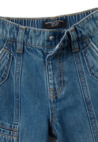 Jeans de mezclilla anchos de color azul claro con una textura desgastada. Presentan trabillas para el cinturón, cuatro bolsillos y costuras en contraste. Etiqueta de la marca visible.