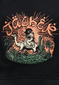 Γυναίκα με πέπλο που οδηγεί μία καμήλα ανάμεσα σε φυτά της ερήμου και κάκτους, με στυλιζαρισμένο κείμενο "Jacquemus" και ηλιαχτίδα στο φόντο.