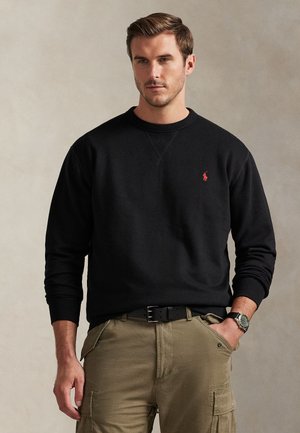 Mand iført sort langærmet pullover med rødt logo, khaki cargobukser, sort bælte og armbåndsur, står med den ene hånd i lommen.