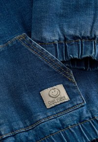 Gros plan sur un tissu en denim bleu avec une poche cousue et une taille élastique, montrant un patch beige avec un visage souriant et le texte "Cool Vibes".