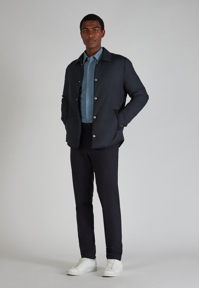Giubbotto imbottito navy con bottoni a pressione argentati, indossato sopra una camicia azzurro chiaro e pantaloni scuri. Abbinato a sneakers bianche.