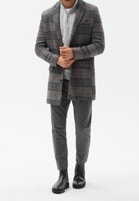 Gråternet frakke med orange accenter, grå lynlås sweater, grå bukser og sorte ankelstøvler, stående mod en almindelig hvid baggrund.