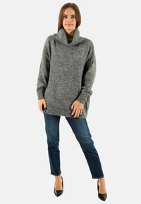 Pull en maille gris avec un large col roulé, des manches longues et une coupe ample, associé à un jean bleu foncé et des escarpins noirs à bout pointu.