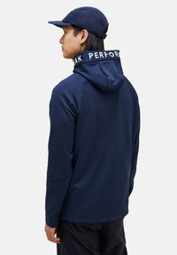 Sudadera de rendimiento azul marino con capucha ajustable, que presenta un cuello alto y el texto blanco "PEAK PERFORMANCE" a lo largo del cuello. Textura suave.
