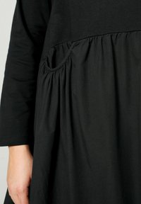 Černé šaty s nařaseným pasem, 3/4 rukávy a kulatou kapsou. Měkká látka s hladkým povrchem. Jednoduchý, uvolněný silueta.