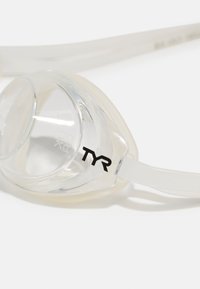 Lunettes de natation transparentes avec un joint en silicone doux et des sangles blanches réglables. Présente le logo noir "TYR" d'un côté.
