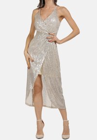 Femme portant une robe portefeuille sans manches en sequins argentés avec une fente sur le côté et des talons beiges à bride de cheville, posant avec une main sur la hanche devant un fond blanc.