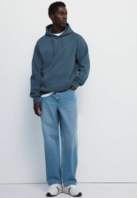 Sweatshirt à capuche bleu foncé avec une poche avant, associé à un jean taille haute bleu clair et des baskets blanches. Texture de tissu lisse.