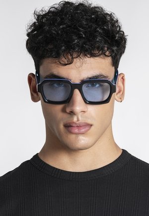 Ung mand med krøllet sort hår iført mørke rektangulære solbriller, lille guldring i øret og en sort ribbet skjorte.