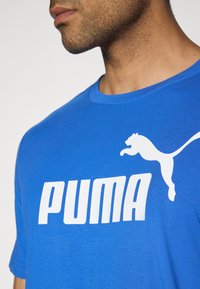 Zila kokvilnas T-kreklā ar balto "PUMA" logotipu un lēcēja pumux grafiku. Īsas piedurknes un apļa kakla dizains.