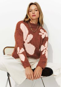 Pull en maille marron avec de grands motifs floraux roses, col montant, poignets côtelés, associé à un pantalon large de couleur claire et des bottes sombres.