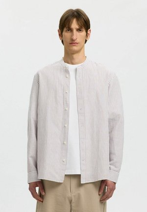 Ung mand iført beige, stribet skjorte med knapper over hvid t-shirt og beige bukser, stående mod en ensfarvet lys baggrund.