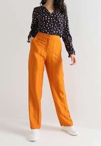 Blusa nera con motivo floreale rosa, abbinata a pantaloni arancioni a vita alta e plissettati. La modella indossa sneakers bianche, mostrando un outfit casual.