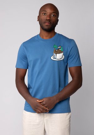 Blaues Baumwoll-T-Shirt mit einem Taschenmotiv, das eine Tasse, einen Berg, Palmen und die Sonne zeigt. Kurze Ärmel und Rundhalsausschnitt.