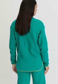 Pullover in fleece turchese con maniche lunghe, orlo arrotondato e piping giallo a contrasto lungo i bordi. Texture morbida e vestibilità comoda.