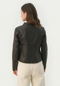 Giubbotto di pelle nera con design aderente, colletto rialzato e polsi con zip. Texture liscia con dettagli di cucitura visibili.