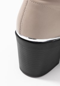 Tacón de zapato de piel beige con costuras visibles, un brillante ribete plateado y un grueso tacón negro en bloque con un patrón texturizado.