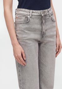 Gros plan d'une personne portant un jean en denim gris clair taille haute avec un bouton et une fermeture éclair, et un haut bleu marine foncé.