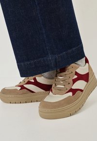 Zapatillas beige y burdeos con materiales de ante y cuero, que presentan una suela de goma gruesa y acentos texturizados, combinadas con unos vaqueros azul oscuro.