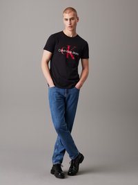Black T-shirt with red logo "Calvin Klein Jeans," paired with blue denim jeans and black leather shoes. Simple design, fitted shape.