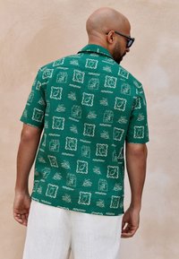 Short-sleeve green shirt featuring a white leaf and frame pattern, made of textured fabric, paired with light-colored linen pants.