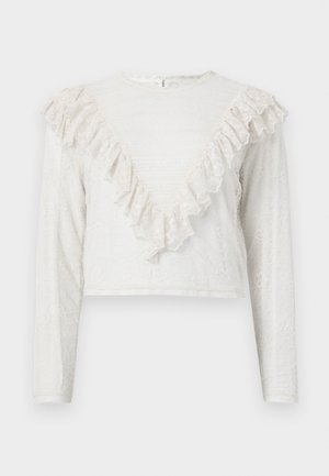 Frau trägt eine weiße Spitzenbluse mit langen Ärmeln und Rüschen, korallenfarbene gemusterte Shorts, schwarze Socken, rote Ballerinas und hält eine kleine weiße Clutch.