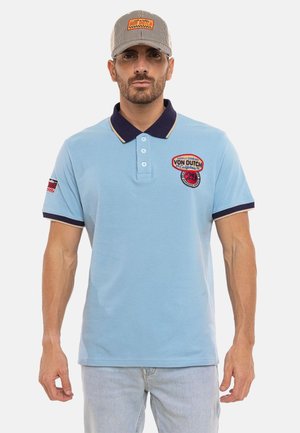 Homme portant un polo bleu clair avec col sombre et écussons, casquette Von Dutch rayée claire, debout sur fond blanc.