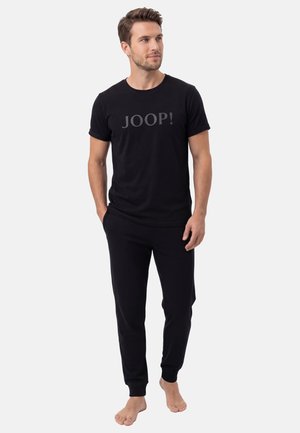 Mężczyzna stojący boso, ubrany w czarny krótki t-shirt z napisem „JOOP!” i pasujące czarne spodnie na jednolitym tle.