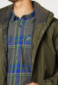 Veste vert olive avec fermeture éclair à l'avant, associée à une chemise en flanelle à carreaux gris et bleu, dotée de deux poches poitrine et d'une fermeture à boutons.