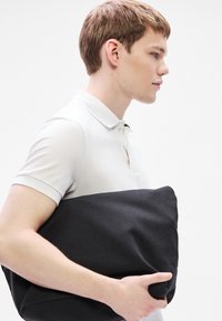 Μαύρη υφασμάτινη τσάντα που κρατιέται κάτω από το μπράτσο από ένα άτομο, με απαλή, υφή επιφάνεια. Το άτομο φοράει λευκό πουκάμισο με γιακά.