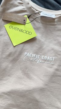 Svijetlosmeđa organska pamučna majica s kratkim rukavima s natpisom "PACIFIC COAST California" izvezenim bijelom bojom. Oznaka je pričvršćena s naglasima u smaragdnozelenoj i smeđoj boji.