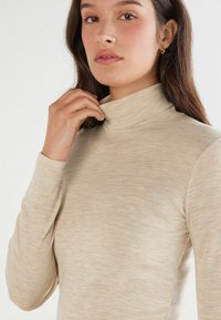 Beige Rollkragenoberteil aus einem weichen, strukturierten Stoff, mit einer taillierten Silhouette und langen Ärmeln. Minimalistisches Design mit einem umgeschlagenen Kragen.
