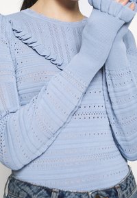 Helles blaues Langarmshirt mit rüschendem Ausschnitt, strukturiertem Strickmuster, Rippbündchen und offenen Webdetails. Getragen mit hochtaillierten Jeans.