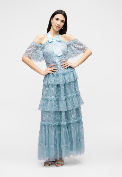 Γυναίκα ποζάρει με ένα ανοιχτό γαλάζιο δαντελένιο φόρεμα με στρωματοποιημένα βολάν και φιόγκο στο ντεκολτέ, φορώντας πέδιλα με ανοιχτά δάχτυλα.