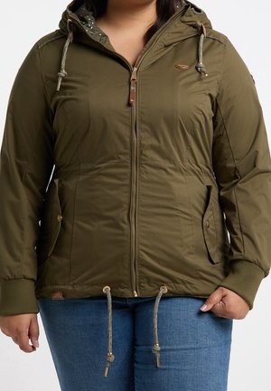 Kobieta ubrana w oliwkowozieloną kurtkę z kapturem na zamek i sznurkami oraz niebieskie dżinsy, dłonie swobodnie opuszczone wzdłuż ciała.