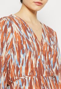 Vestido marrón y crema con acentos azules; presenta un escote en V, mangas largas y un diseño fluido con un patrón que recuerda a pinceladas.