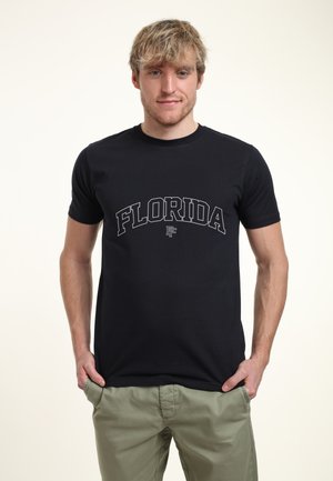Ung mand med rodet blondt hår iført en sort t-shirt med teksten "FLORIDA" og lys grønne bukser, stående med hænderne i lommerne.