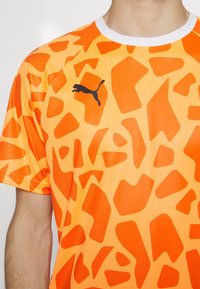 Camiseta corta de manga naranja con un diseño de patrón de jirafa con formas variadas y un logotipo negro de Puma en el pecho. Con ribete blanco en el cuello.