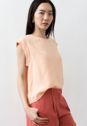 Kobieta o długich ciemnych włosach ubrana w jasnoróżową, bezrękawnikową bluzkę i wysokie, rdzawo-brązowe spodnie, stojąca na jasnym tle.