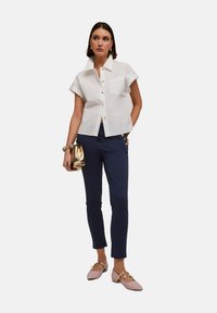 Camisa blanca de botones de manga corta con un bolsillo, combinada con pantalones ajustados azul marino con acentos de botones dorados y bailarinas rosas claras.