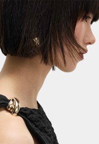 Czarna, strukturalna tkanina opada na ramię, z wyrazistym, metalicznym złotym akcentem. Krótkie, ciemne włosy okalają twarz.