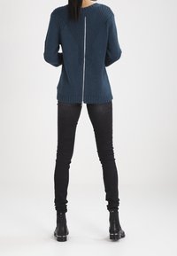 Pull tricoté bleu sarcelle avec un motif en V dans le dos, équipé d'une fermeture éclair centrale. Associé à un jean noir slim et des bottines.