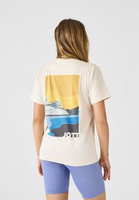 Camiseta color crema con un gráfico de paisaje en amarillo, azul y negro en la parte posterior. Mangas cortas y corte relajado.
