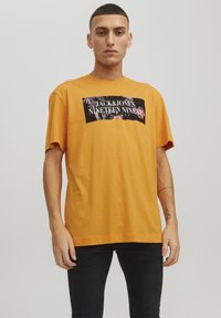 T-shirt di cotone arancione con grafica nera e dettagli floreali e testo "JACK & JONES NINETEEN NINETY." Vestibilità rilassata, maniche corte.
