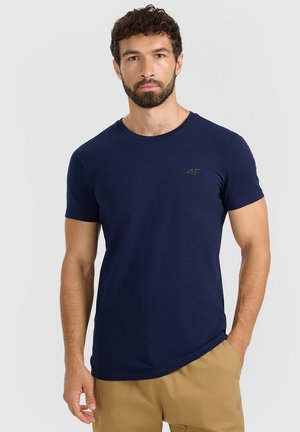 Mężczyzna z kręconymi ciemnymi włosami i brodą, ubrany w granatowy t-shirt i beżowe spodnie, stoi z jedną ręką w kieszeni.