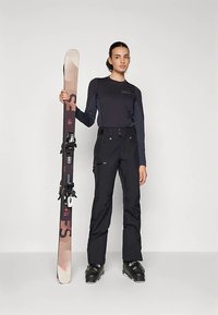 Persona, kas valkā melnu niezi ar garām piedurknēm un melnas slēpošanas bikses, tur rokās brūnu un bēša krāsas snovbordu ar stiprinājumiem. Gludi audumi un mūsdienīgs dizains.