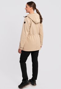 Beige softshell jakke med hætte, snoretræk i taljen og sort accentpatch. Parret med sorte bukser og robuste sorte sko.